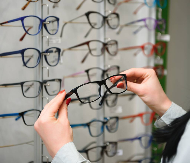 Row of glasses at an opticians. Eyeglasses shop. Stand with glasses in the store of optics. Woman's hand chooses spectacles. Eyesight correction. - Foto, afbeelding