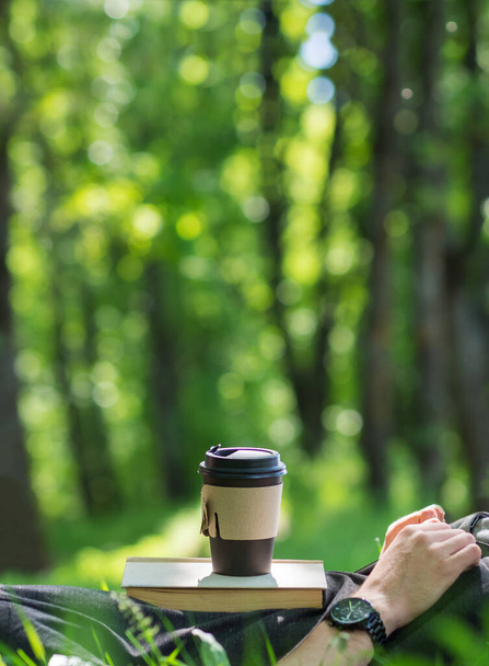 Close-up de uma xícara de papel de café em um livro, um homem deitado na grama verde em um parque em uma manhã de verão - Foto, Imagem