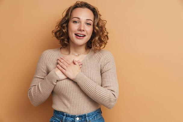 Young ginger woman smiling while holding hands on her chest isolated over beige background - Fotografie, Obrázek