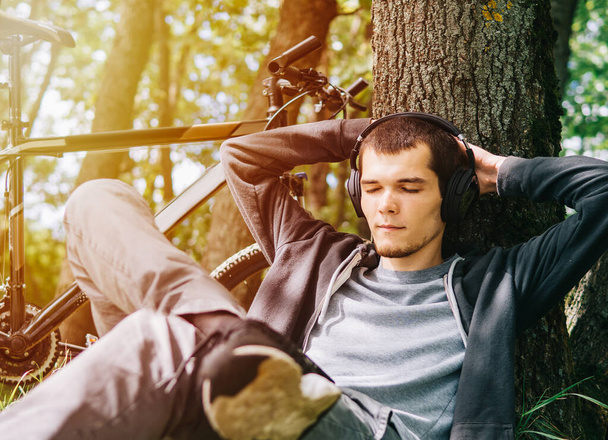 Acuéstese en el parque junto a un árbol y disfrute escuchando música, relájese después de un paseo en bicicleta - Foto, Imagen