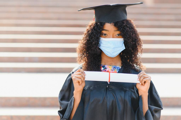 Young female african american graduate student wears a protective mask against coronavirus. Concept of graduation ceremony, quarantine. - Zdjęcie, obraz
