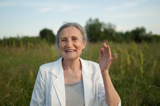 Gri saçlı, yüzü kırışık, beyaz ceketli ve güneşli bir günde parkta dinlenen yaşlı bir kadının portresi. - Fotoğraf, Görsel