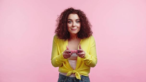 KYIV, UKRAINE - JUNE 30, 2021: young curly woman holding joystick and playing video game isolated on pink  - Foto, Imagen