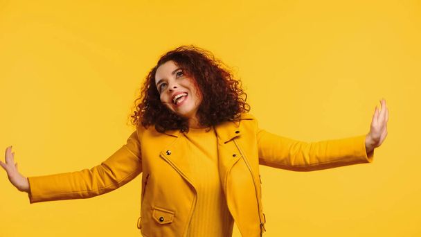 amazed young woman in leather jacket with outstretched hands isolated on yellow - Fotoğraf, Görsel