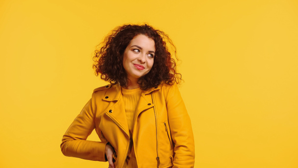 усміхнена молода жінка в шкіряній куртці стоїть з рукою на стегні ізольовано на жовтому
 - Фото, зображення
