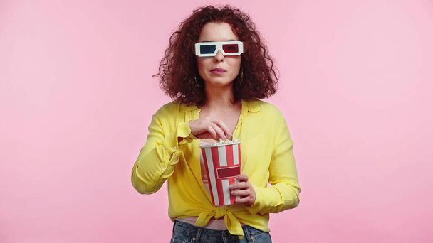 curly young woman in 3d glasses holding popcorn bucket isolated on pink  - Foto, Bild