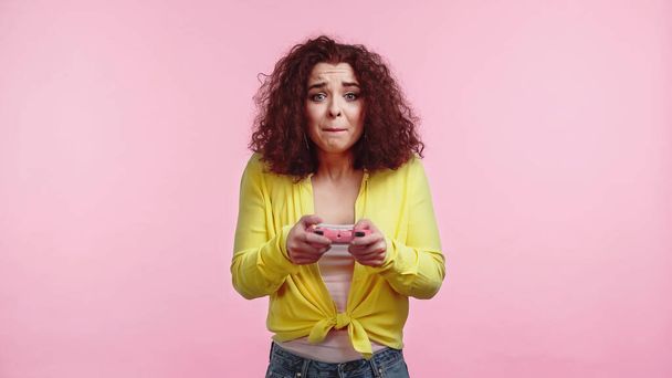 KYIV, UKRAINE - JUNE 30, 2021: worried woman holding joystick and playing video game isolated on pink  - Photo, Image