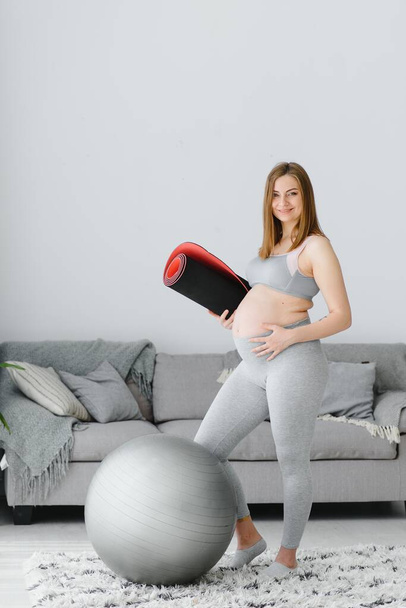 Portrait of a beautiful pregnant woman smiling and holding a rolled yoga mat under her arm. Adorable expectant mother touching her belly and preparing to start her home workout - Fotó, kép
