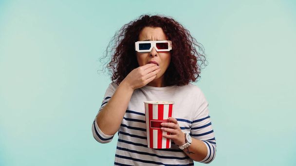 кучерява молода жінка в 3d окулярах їсть попкорн ізольована на синьому
  - Фото, зображення