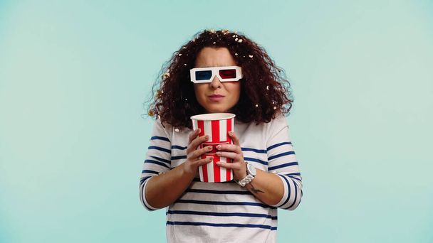 curly young woman in 3d glasses with bucket and popcorn in hair isolated on blue  - Photo, Image