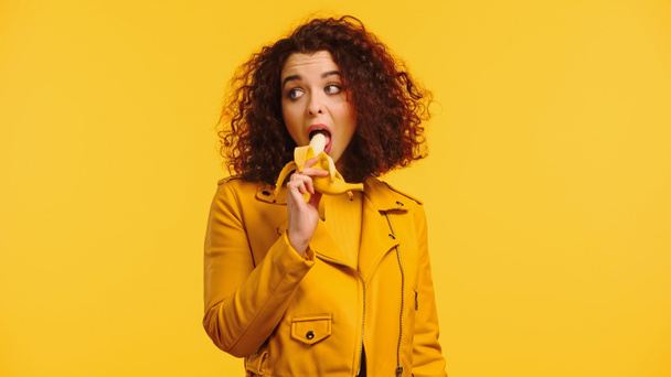 young curly woman in leather jacket eating banana isolated on yellow  - Foto, immagini