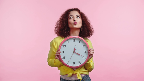 curly woman holding clock and pouting lips isolated on pink  - Fotografie, Obrázek