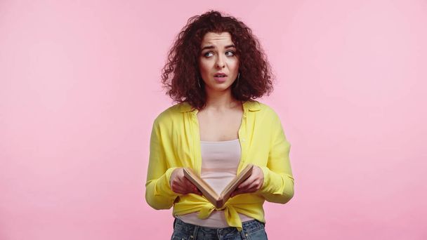 curly and sad woman holding book while studying isolated on pink  - Foto, imagen