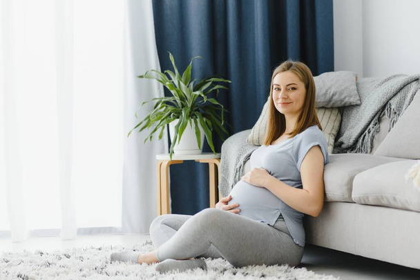 Pregnant woman caressing her belly at home. Young expectant blonde feeling her baby push, sitting on floor, copy space. Pregnancy, rest, life, expectation concept - Фото, изображение