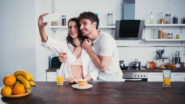 szczęśliwy mężczyzna obejmujący kobietę w rozpiętej koszuli robi selfie podczas śniadania - Zdjęcie, obraz