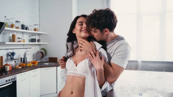 young man embracing sexy, smiling woman in shirt and bra in kitchen - Photo, Image