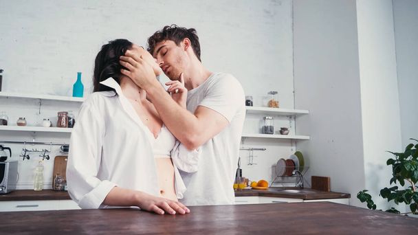 man in white t-shirt kissing seductive woman near table in kitchen - Фото, изображение