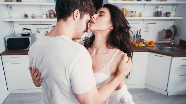 Молодой человек в белой футболке обнимает горячую женщину в лифчике на кухне - Фото, изображение