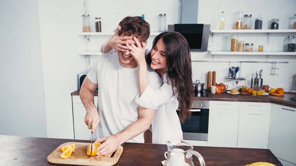 cheerful woman in white shirt covering eyes of man cutting orange in kitchen - Foto, afbeelding