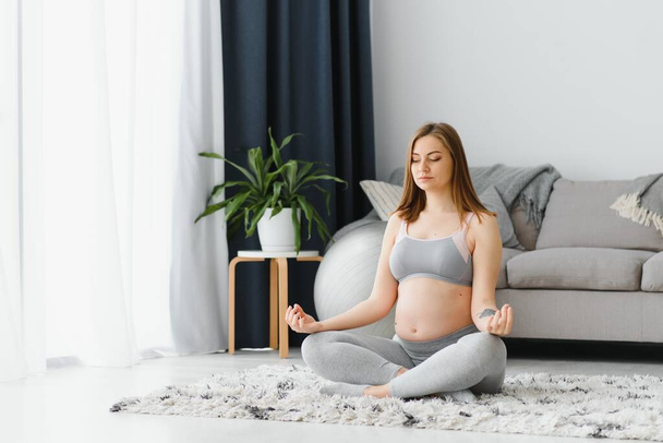 Beautiful pregnant woman sitting on yoga mat in living room, practicing meditation, young expectant lady doing sports at home, enjoying healthy lifestyle during pregnancy time, copy space - Foto, immagini