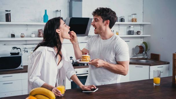 young man with pancakes feeding sexy girlfriend with raspberry in kitchen - 写真・画像