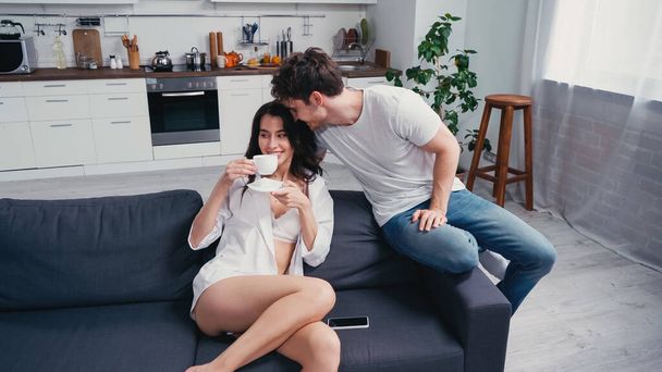hombre feliz cerca de la mujer complacida beber café en el sofá en camisa blanca y sujetador - Foto, imagen