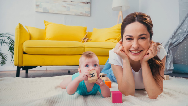glückliche Mutter blickt in die Kamera, während ihr kleiner Sohn mit Spielzeug spielt  - Foto, Bild