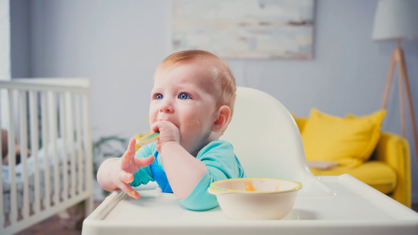 chłopiec z niebieskimi oczami siedzący na krześle do karmienia i jedzący jedzenie dla niemowląt - Zdjęcie, obraz