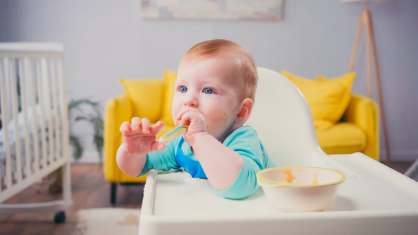 infant boy with blue eyes sitting in feeding chair and sucking spoon near bowl with baby food - Photo, Image