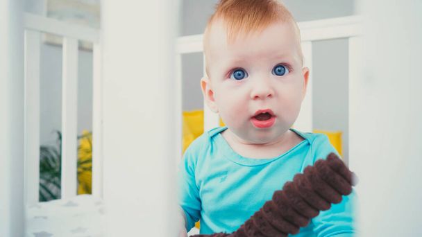 удивленный мальчик с голубыми глазами, сидящий в расплывчатой детской кроватке с мягкой игрушкой - Фото, изображение