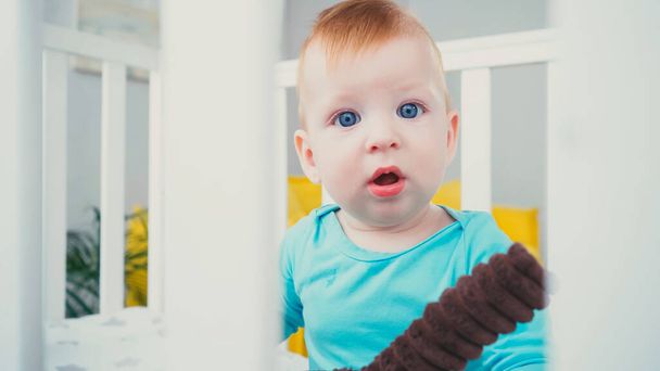 surprised baby boy sitting in blurred baby crib with soft toy - Foto, Imagem