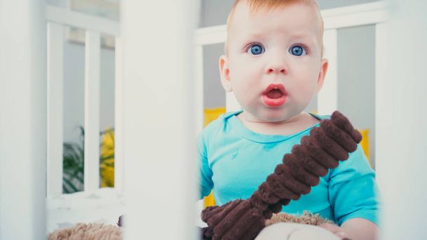 surprised infant boy sitting in blurred baby crib with soft toy - Fotoğraf, Görsel