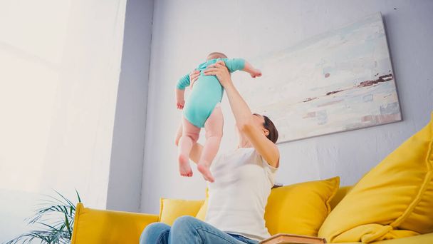 vista a basso angolo di madre che solleva il figlio neonato a piedi nudi mentre è seduto sul divano - Foto, immagini