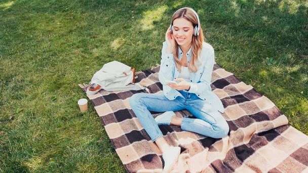 high angle view of happy student in headphones using smartphone on blanket in park - Photo, Image