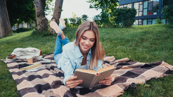 Junge fröhliche Studentin liest Buch, während sie auf karierter Decke im Park liegt  - Foto, Bild