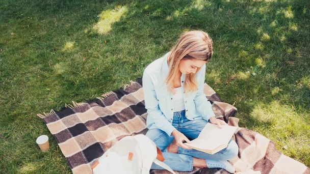 high angle view of student reading book on checkered blanket in park - Foto, Imagen