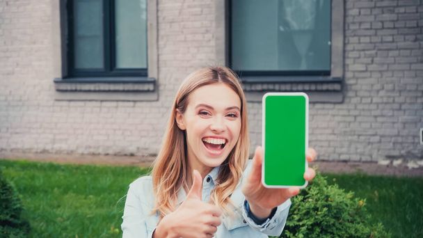 vrolijke student houdt wazig smartphone met groen scherm en duim omhoog in de buurt van gebouw  - Foto, afbeelding