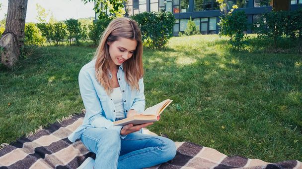 Gülümseyen genç bir kadın, battaniyeye oturmuş parkta kitap okuyor.  - Fotoğraf, Görsel