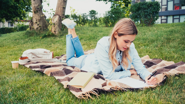 étudiant écrit dans un cahier près de livres tout en étant couché sur une couverture dans le parc - Photo, image