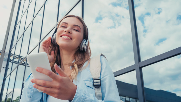 low angle view of cheerful young woman using smartphone and listening music in wireless headphones near building - Valokuva, kuva