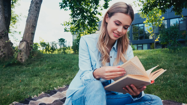 Parkta kareli battaniyenin üzerinde oturmuş kitap okuyan neşeli öğrenci.  - Fotoğraf, Görsel