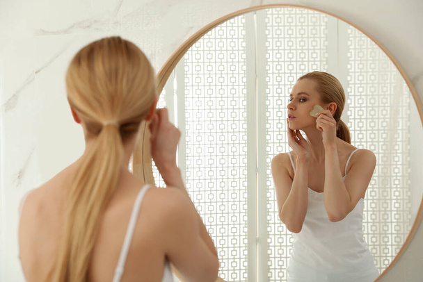 Beautiful young woman doing facial massage with gua sha tool in front of mirror at home - Fotografie, Obrázek
