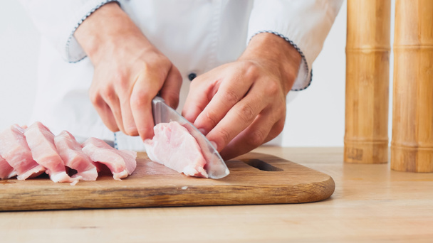 vista parcial del cocinero rebanando lomo de cerdo sobre tabla de cortar en blanco - Foto, Imagen