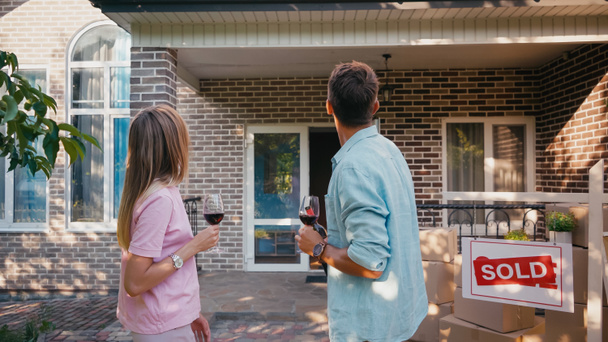 back view of man and woman holding glasses of red wine near board with sold lettering and new house - Photo, Image