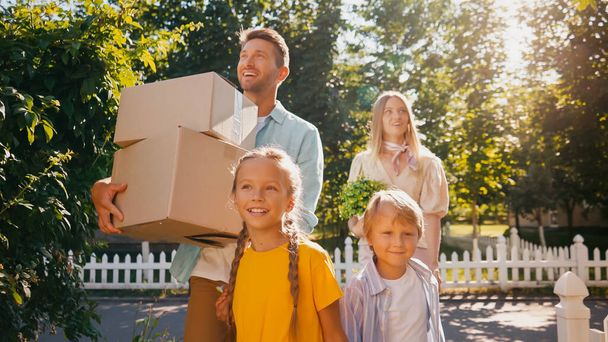 cheerful parents holding boxes and plant while walking with happy kids, relocation concept - Fotoğraf, Görsel