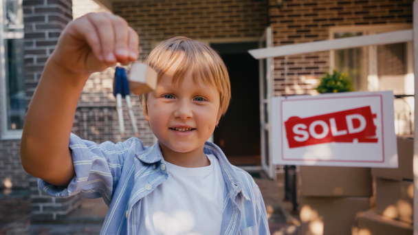 joyful boy looking at camera and holding keys near new house - Photo, Image