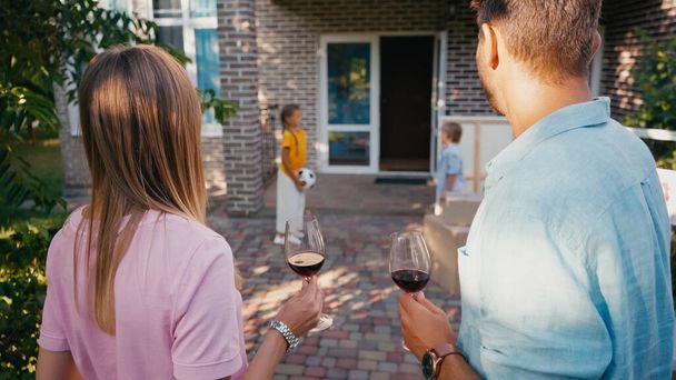 couple holding glasses of wine near blurred kids and new house - Foto, Imagen