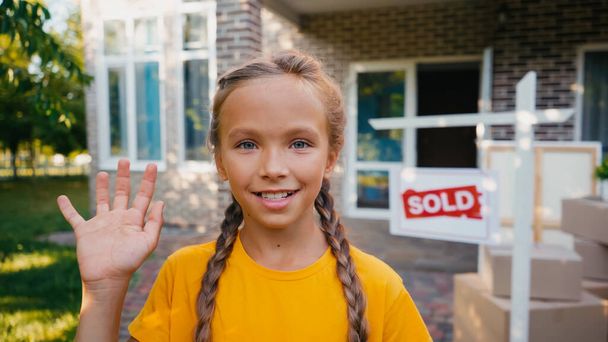 joyful kid looking at camera and waving hand near blurred board with sold lettering and new house - Photo, Image