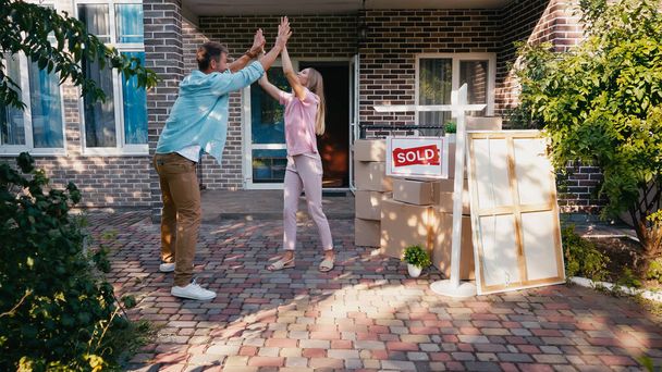 joyful couple giving high five near new house and sold board  - Photo, image