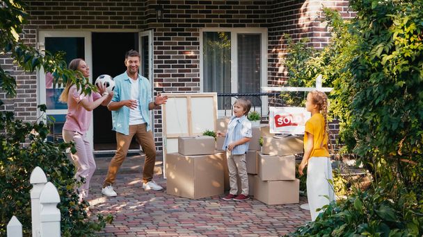 madre lanzando fútbol a los niños cerca de marido, cajas de cartón y casa nueva - Foto, Imagen
